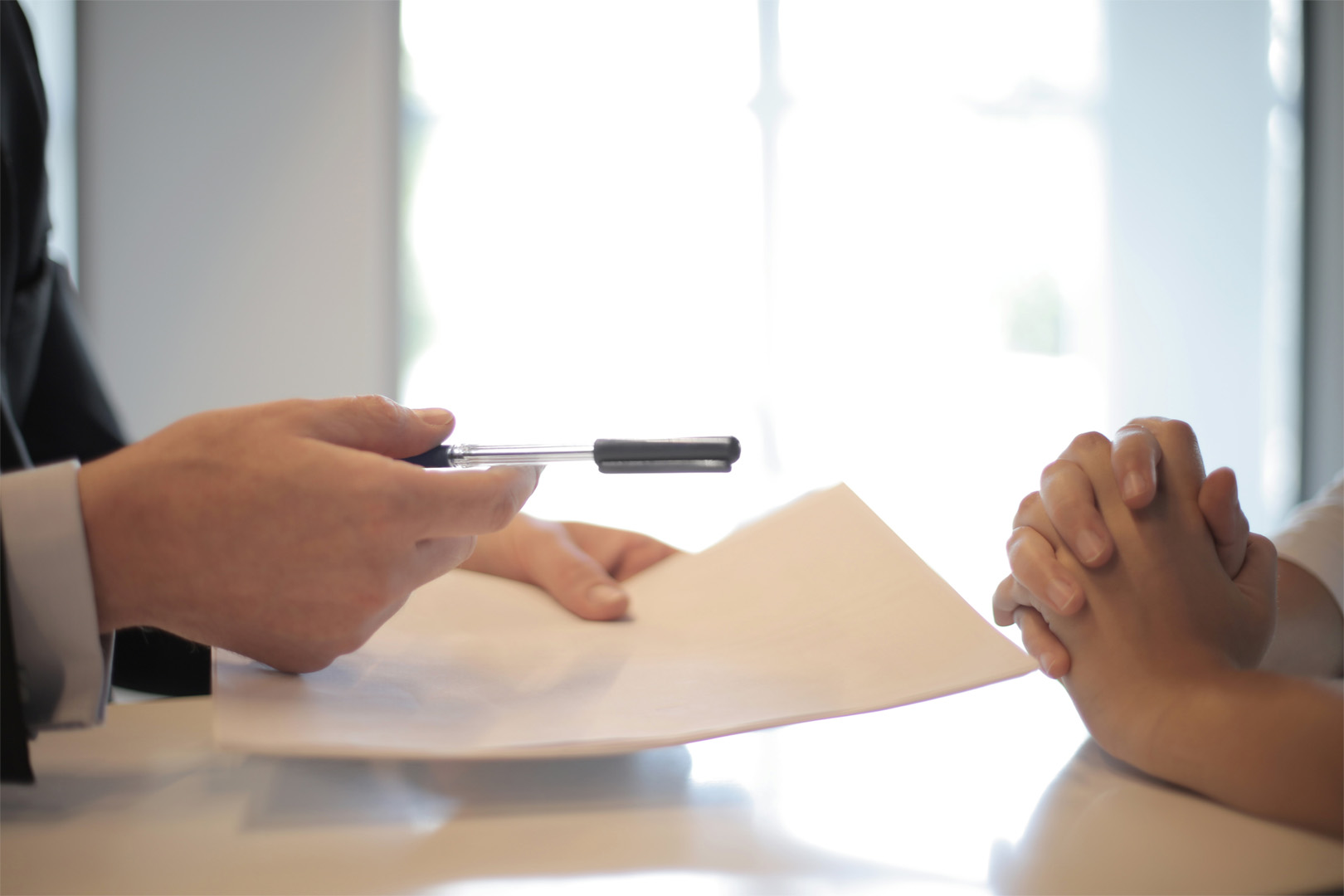 Close-up shot of someone holding a pen and documents in their hands, opposite an individual with clasped hands in front of their body on a table