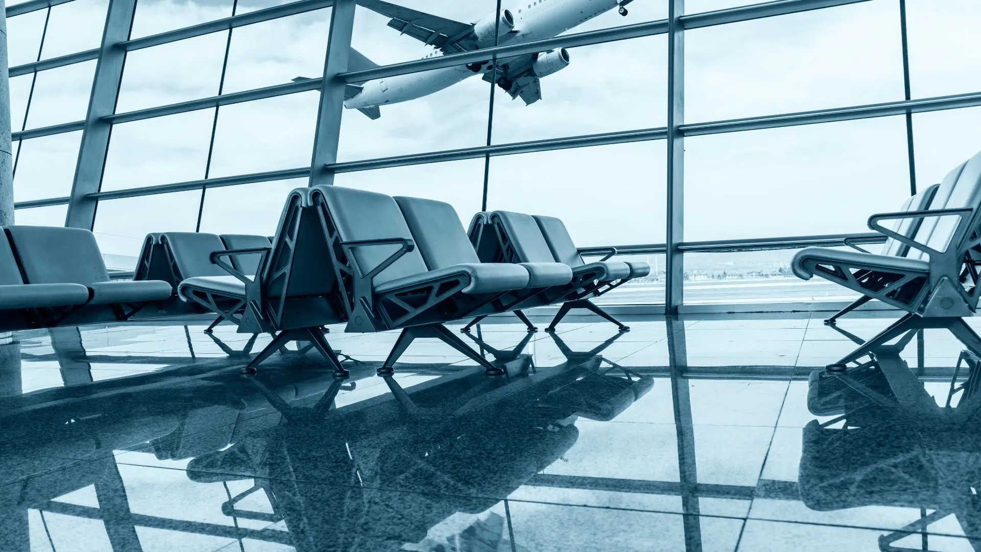 Blue-tinged shot of an airport waiting area, featuring a seated area overlooking the windows in the background of the shot, an airplane is taking off in the background