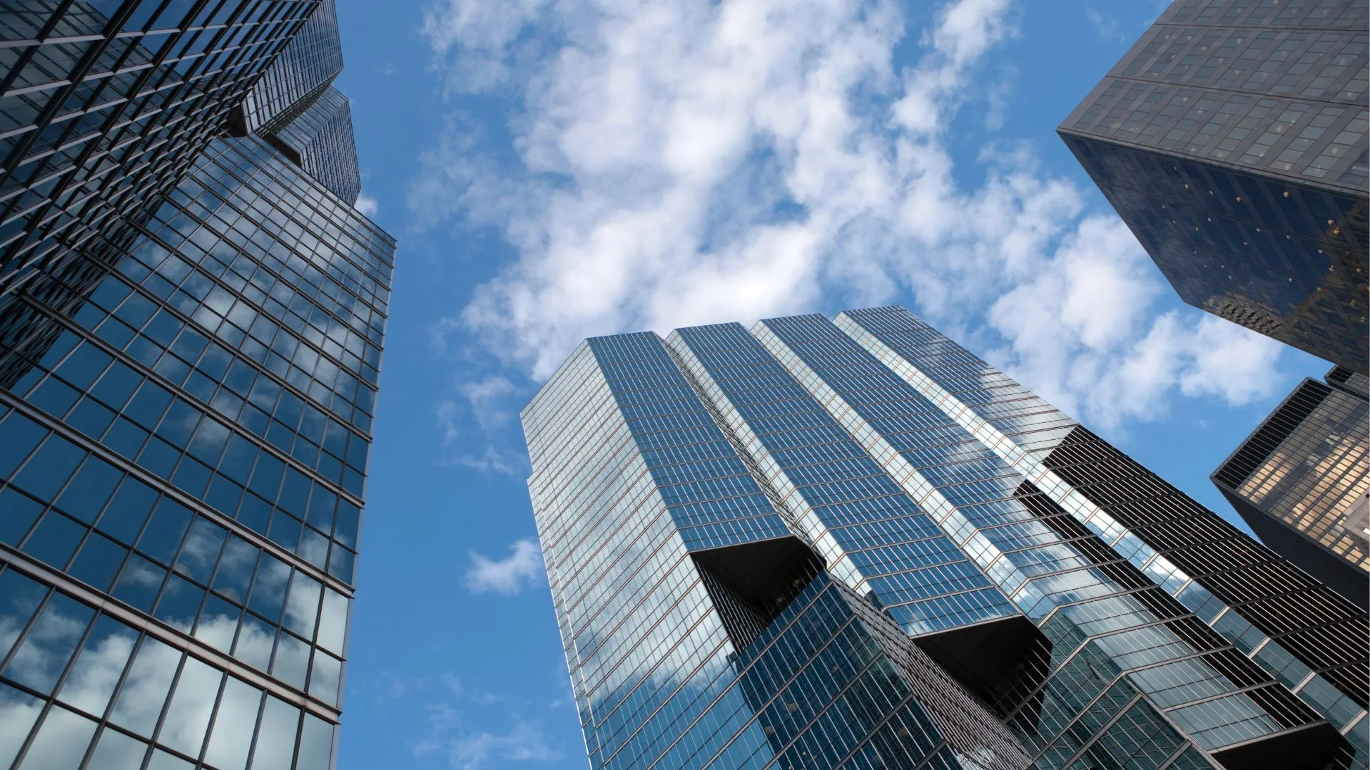 Shot of multiple glass-fronted skyscrapers shot from the ground-up, a bright blue sky with some clouds can be seen above