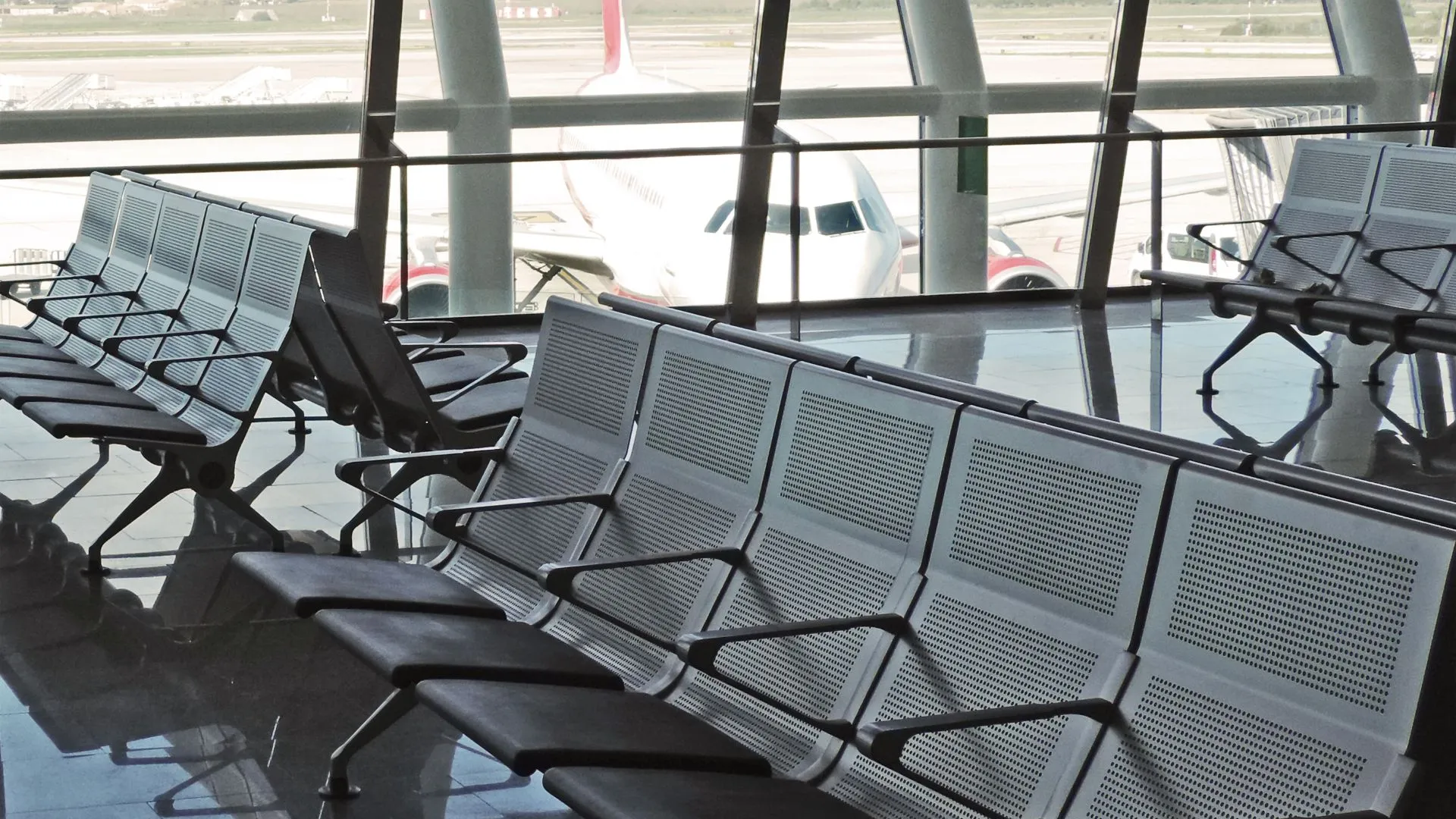 Shot of an airport waiting area, featuring a seated area overlooking the windows in the background of the shot, an airplane is grounded in the background outside