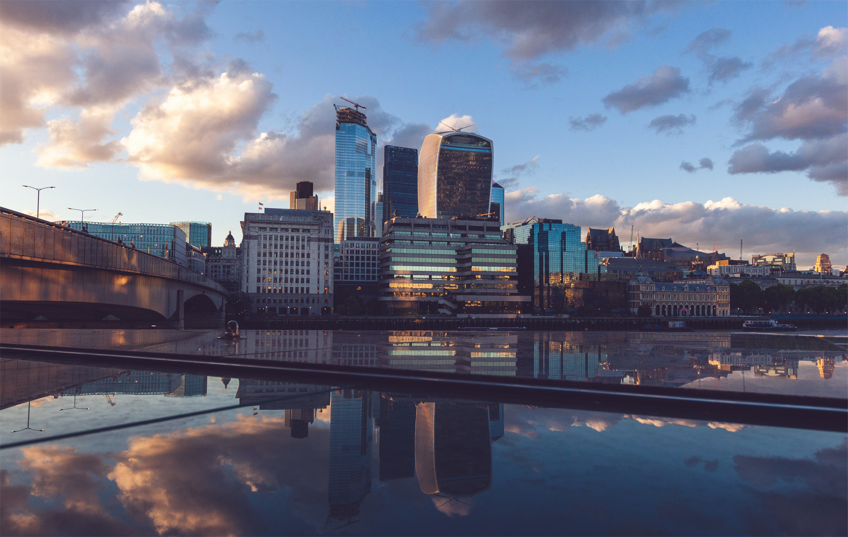 Landscape shot over a river in the foreground featuring a city scape in the background of the shot, including multiple buildings of various heights and aesthetics, some more traditional and some more modern. A bridge crossing the river can be seen in the left-hand side of the frame.