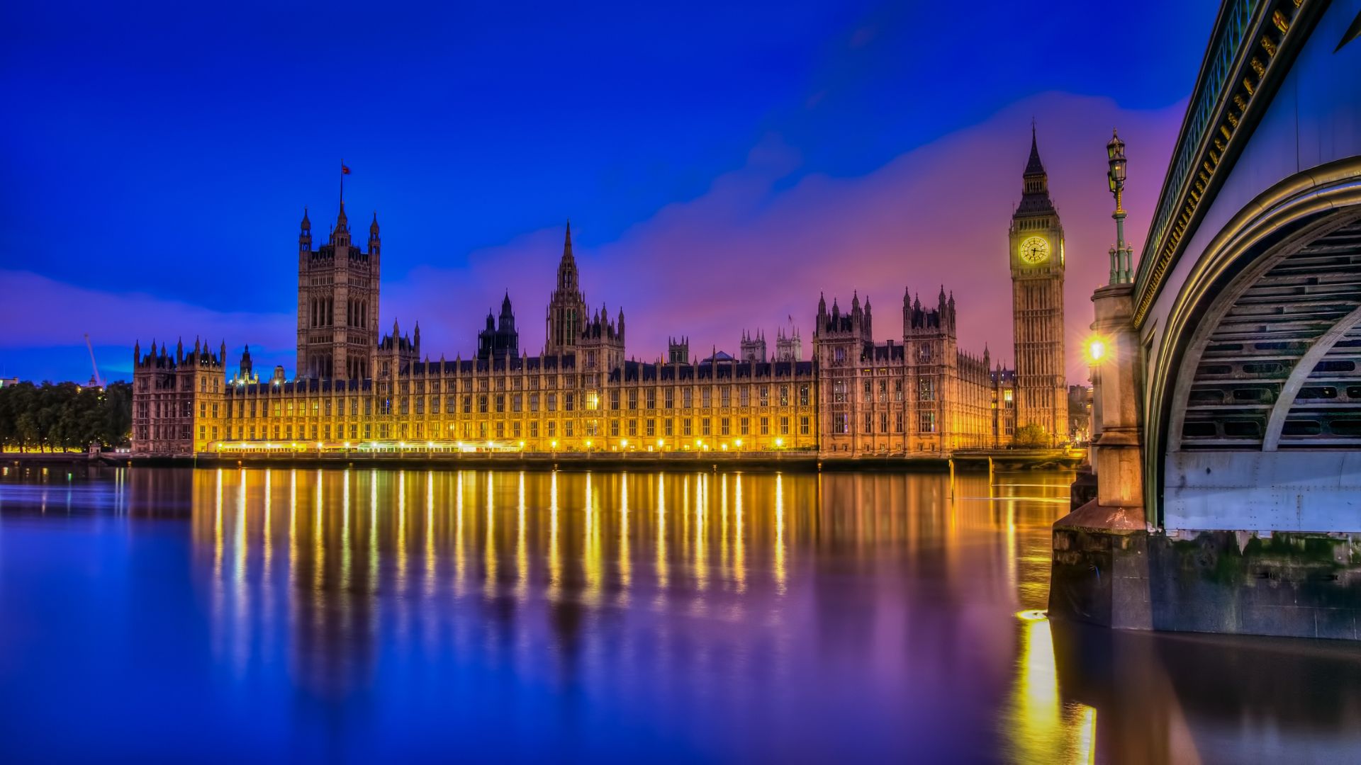 Shot of the Houses of Parliament in London, UK at night, the sky is purple and dark blue, as is the River Thames in the foreground of the shot.